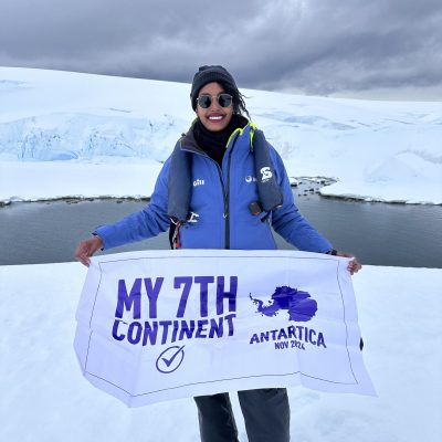A Black woman traveling alone in Antarctica holding a flag that reads "My 7th Continent" during her solo journey.